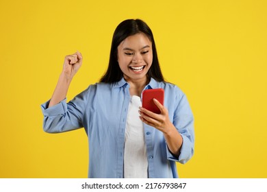 Glad Excited Young Chinese Lady Looking At Smartphone And Making Victory Gesture With Hand Isolated On Yellow Background, Studio. Online Victory, New App, Shocking Ad And Offer, Great News And Tech