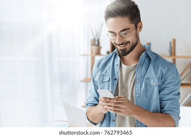 Glad fashionable young bearded guy wears stylish clothes and denim shirt, has trendy hairstyle, happy to exchange messages with friends, uses free internet connection on electronic modern gadget
