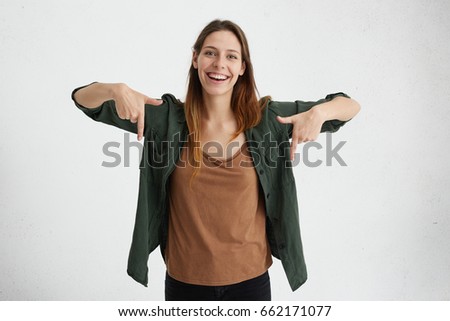Glad female with oval face, dark straight hair wearing green jacket and brown shirt pointing with her index fingers down having cheerful look while advertising something. Advertising concept