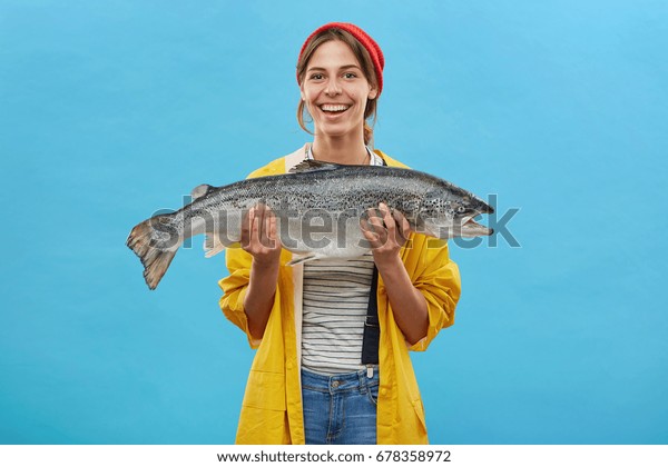 Glad fisher woman in yellow anorak and jeand\
overalls holding huge fish rejoicing to catch it demonstrating her\
work while standing over blue background. People, hobby, recreation\
and fishing