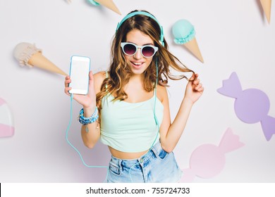 Glad girl in blue accessories listening music in her room, holding smartphone and playing with hair. Portrait of elegant young woman in glasses with curly amazing hairstyle posing on purple wall.