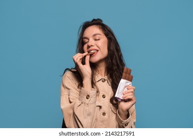 Glad happy millennial european woman with closed eyes enjoys taste of chocolate bar, isolated on blue background, studio shot, free space. Sweet tooth, love of sweets and candy, ad and good offer - Powered by Shutterstock