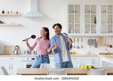 Glad Happy Young European Man And Woman Have Fun Together, Singing Song At Imaginary Microphone, Preparing Food In Kitchen Interior. Couple Enjoy Cooking, Positive Mood And Superstar, Covid-19 Concept