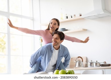 Glad Happy Young Husband Carries His Wife On Back, Have Fun Together On Kitchen Internet. Couple Enjoy Free Time Together In Own New Home. Great Offer, Ad, House And Life Insurance, Empty Space