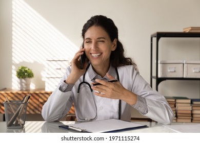 Glad To Hear You Feel Well. Happy Young Latin Lady Doctor Sit At Desk Talk To Patient By Phone Enjoy Listening About Recovery Improving Health. Positive Woman Medic Nurse Speaking By Cell From Cabinet