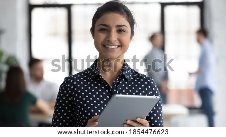 Glad to help you! Portrait of smiling confident indian female insurance broker bank manager hr assistant standing in open space office holding digital tablet looking at camera ready to assist client