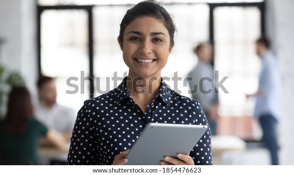 Glad to help you! Portrait of smiling confident
indian female insurance broker bank manager hr assistant standing
in open space office holding digital tablet looking at camera ready
to assist client