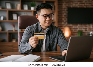 Glad Mature Asian Man In Glasses Typing On Laptop, Showing Credit Card At Table In Home Office Interior. Finance Manager, Freelance, Businessman Shopping Online, Enjoy Sale And Check Money On Account