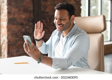 Glad To Meet. Cheerful Happy Indian Man In Casual Wear Using Smartphone App For Virtual Communication, Holding Phone In Hand And Waving At Webcam, Holding Video Call Sitting At The Desk