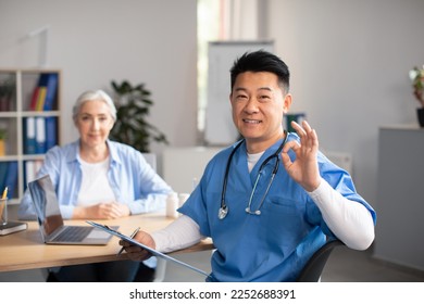 Glad middle aged korean doctor showing ok sign with hand, approving treatment result of old european female patient in clinic office interior. Recommendation and advice from therapist, health care