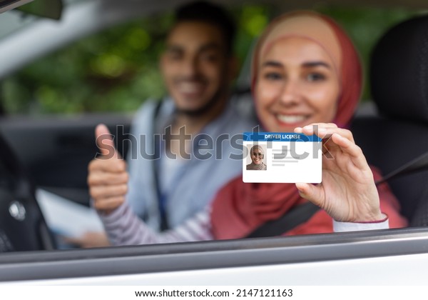 Glad middle eastern male and female in hijab\
show driving license and thumb up in open window in car after exam,\
outdoor in summer. Get license, driving test, lesson with teacher\
and life insurance