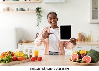 Glad millennial black female pointing finger at tablet with blank screen at table with organic fruits and vegetables in scandinavian kitchen interior. Food blog, health care at home and app for work
