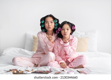 Glad millennial japanese lady and teen girl in pajamas and curlers with cosmetics on bed have fun and make air kiss in bedroom interior. Mom and daughter do skin treatments and makeup, beauty day
