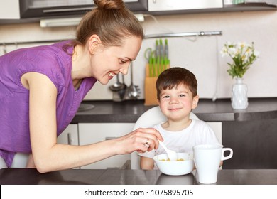 Glad mother feeds her little male child with spoon, gives delicious porridge and tea, promises to go for walk after dinner, sit at kitchen interrior. Smiling young woman prepares cereals for kid.