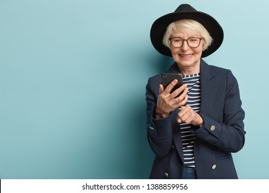 Glad Old Woman Receives Good News On Smart Phone, Types Feedback, Wears Fashionable Black Hat, Jacket And Striped Jumper, Happy To Learn Using Modern Technologies, Stands Indoor Over Blue Wall