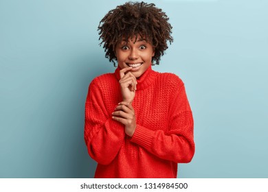 Glad pleased dark skinned young woman with crisp hair, keeps hand near mouth, wears red jumper, looks happily, listens something positive with great interest, stands against blue background. - Shutterstock ID 1314984500