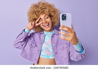 Glad positive European woman with curly blonde hair makes peace gesture over eye smiles happily takes selfie via smartphone wears stylish clothes poses against purple background enjoys life.