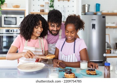Glad positive young family foolishes with small daughter at kitchen, making pancakes, have tasty breakfast, pose at camera, drink fresh milk, eat pancakes
