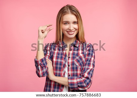 Glad pretty woman shows something tiny with hands, wears checkered shirt, glad to play with little kid, isolated over studio pink background. Beautiful young female demonstrates size of heel