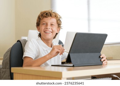 Glad schoolboy using stylus on tablet during lesson while sitting at desk in classroom at school and looking forward - Powered by Shutterstock