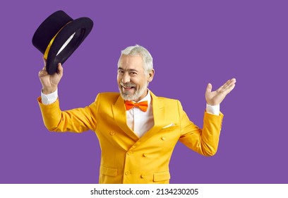 Glad to see you. Joyful positive presenter greeting you at his show. Happy polite senior man wearing stylish bright yellow suit and orange bow tie looks at camera, takes off black top hat and smiles