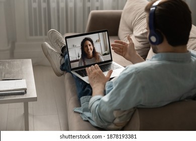 Glad To See You. Over The Shoulder View Of Peaceful Millennial Man Relaxing On Sofa In Earphone Set With Computer On Laps. Calm Young Husband Chatting With Wife By Video Link Using Headphones And Pc