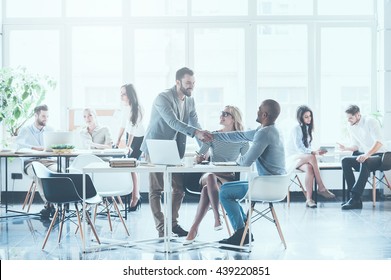 Glad To See You In Team! Group Of Young Business People Working And Communicating With Each Other In Office While Two Men Shaking Hands And Smiling