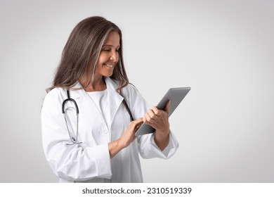 Glad senior doctor woman in white coat with stethoscope using digital tablet on gray studio background, free space. Medical care remotely, diagnostics, ad and offer - Shutterstock ID 2310519339