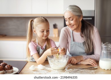 Glad Smiling Little Girl And Senior Woman In Aprons Make Dough, Break Egg In Minimalist Kitchen Interior. Cooking Lesson Baking, Household Chores Together, Prepare Food For Family At Home Due Covid-19