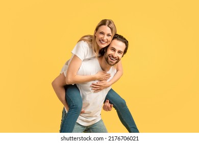 Glad smiling millennial caucasian woman on man on back in white t-shirt, enjoy moment, have fun at free time, isolated on yellow background, empty space. Ad and offer, couple relationship and love