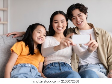 Glad Smiling Young Asian Couple And Teen Daughter Making Photo On Phone On Sofa In Living Room Interior. Covid-19 Quarantine, Social Distance, Video Call, Chat And Family Selfie At Home Due Covid-19