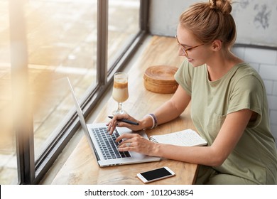 Glad successful female freelancer with hair knot provides online marketing service, surves websites and social networks, promotes some products, works during free time at cafe with smoothie.