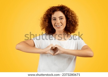Glad teen woman forming heart sign with her hands on chest, isolated on yellow background, exemplifying cheerful, love filled expression and youthful vibrancy in lively, positive, sunny setting