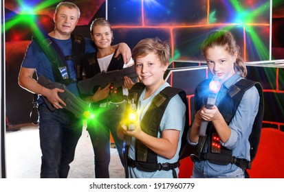 Glad teenage boy and girl posing with laser guns while having fun with adults on laser tag arena
