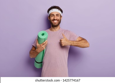 Glad Unshaven Man Makes Okay Gesture, Keeps Thumb Up, Holds Rolled Up Karemat, Shows He Liked Yoga Training Wears Casual T Shirt In One Tone With Background. People, Workout, Healthy Lifestyle Concept