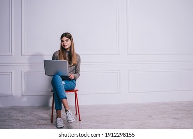 Glad woman freelancer looking at camera sitting on chair with crossed legs and laptop on lap while doing work task