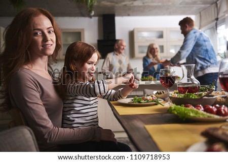 Glad woman holding daughter on lap. They are sitting while rest of family members bringing dishes to table on background. Mother is smiling while her kid is teasing