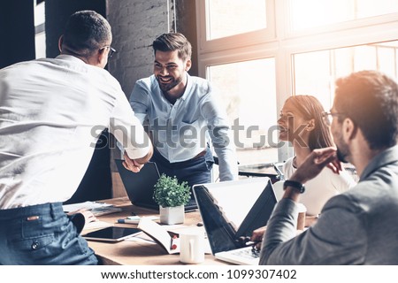 Glad to work with you! Young modern men in smart casual wear shaking hands and smiling while working in the creative office