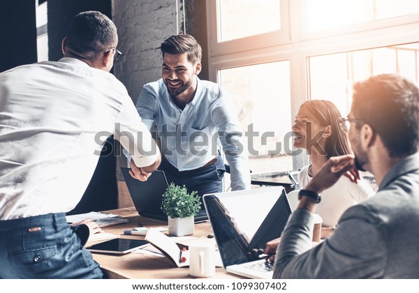 Glad to work with you! Young modern men in smart\
casual wear shaking hands and smiling while working in the creative\
office
