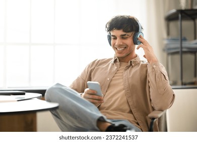 Glad Young Arab Guy Having Fun With Smartphone, Listens Online Music In Wireless Earphones, Sitting Near Desk During Work Day At Home Interior. Modern Gadgets Lifestyle Concept - Powered by Shutterstock