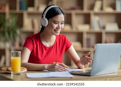 Glad young asian lady student in wireless headphones with juice looking at laptop in living room interior. Online call, education and study at home, video lesson, social distance, covid-19 quarantine