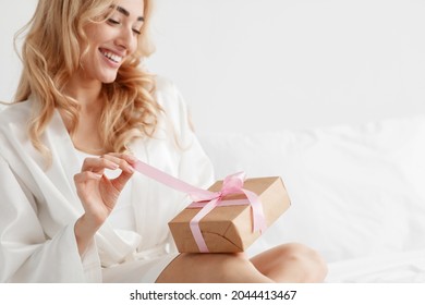 Glad young attractive european blonde female in robe, opens gift box, unties ribbon on white bed in bedroom, close up, copy space. Anniversary, happy birthday, Valentine day and positive emotions - Shutterstock ID 2044413467