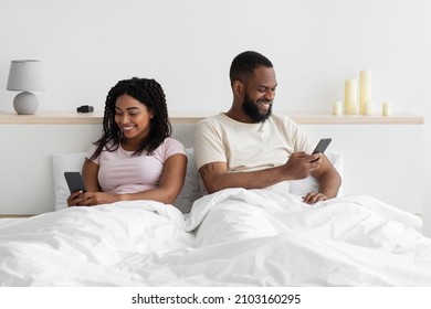 Glad young black female ignores her husband sitting on bed and look at phone in bedroom interior, copy space. Treason, addiction on social networks and technology, personal information and new normal