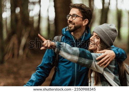 Glad young caucasian couple in jackets enjoy journey, points finger at copy space, rest in forest, outdoor, close up. Hiking, active lifestyle, adventure and tourism, people emotions