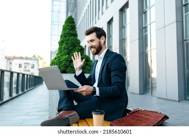 Glad Young European Businessman With Beard In Suit Has Video Call On Laptop, Waving Hand Near Modern Office Building, Outdoor. Business Meeting Remote, Coffee Break, Social Distance And New Normal