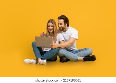 Glad Young European Man With Beard And Woman Hugs, Sit On Floor, Look At Laptop, Isolated On Yellow Background, Studio. Modern Technology, Relationship, Video Call, Meeting Remotely And New Normal