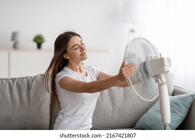 Glad Young European Woman Suffers From Heat Sits On Sofa Catches Cold Air, Turns Fan To Herself, Enjoys Cool Down In Living Room Interior. Summer Weather, Overheating Without Air Conditioning At Home
