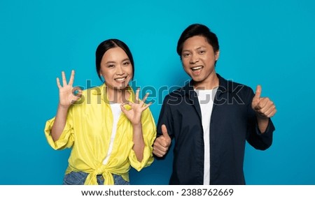 Glad young japanese man and lady in casual show ok, thumb up sign, celebrate win, success with hand, isolated on blue studio background. Emotions recommendation, approve gesture, good news sign