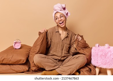 Glad young woman sits in lotus pose on soft bed wears pajama wrapped towel on head laughs happily away feels relaxed after sleeping covered with blanket surrounded by pillows and alarm clock