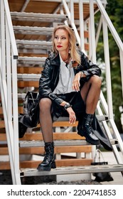 Glam Rock Style In Clothes. A Woman In A Black Leather Coat, Black High Boots With Spikes, Chains Around Her Neck. Fishnet Tights With Black Leather Shorts.